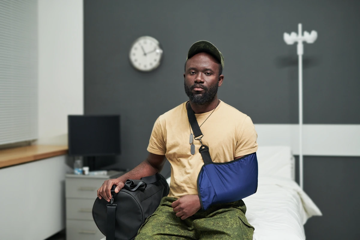 Un homme assis sur un lit d'hôpital, le bras dans une écharpe, avec sa valise